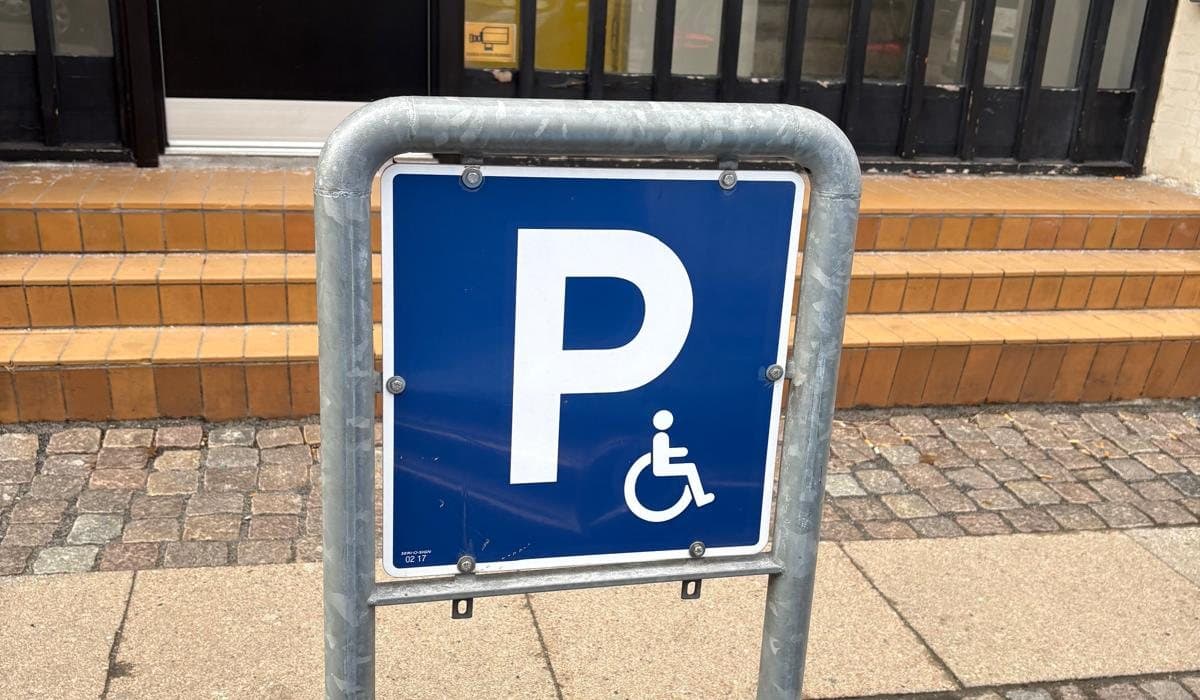 Blåt handicapparkeringsskilt med kørestolssymbol i et galvaniseret skiltestativ på et fortov.