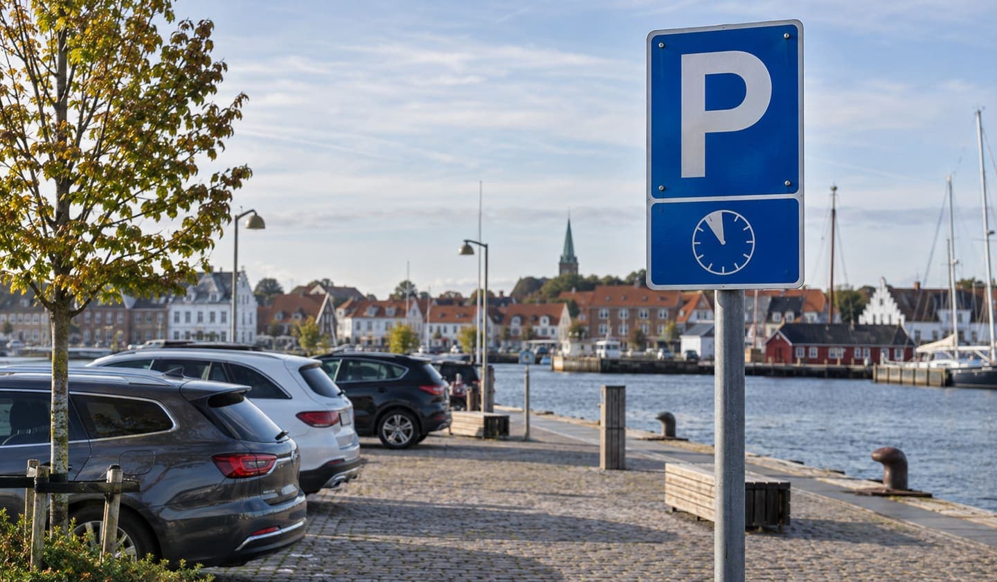Parkeringsskilt ved en dansk havneplads med biler, vand og byhuse i baggrunden.