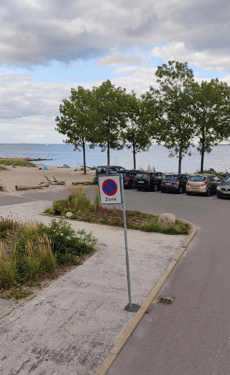 Parkerede biler ved et kystområde med et zonenskilt i forgrunden.