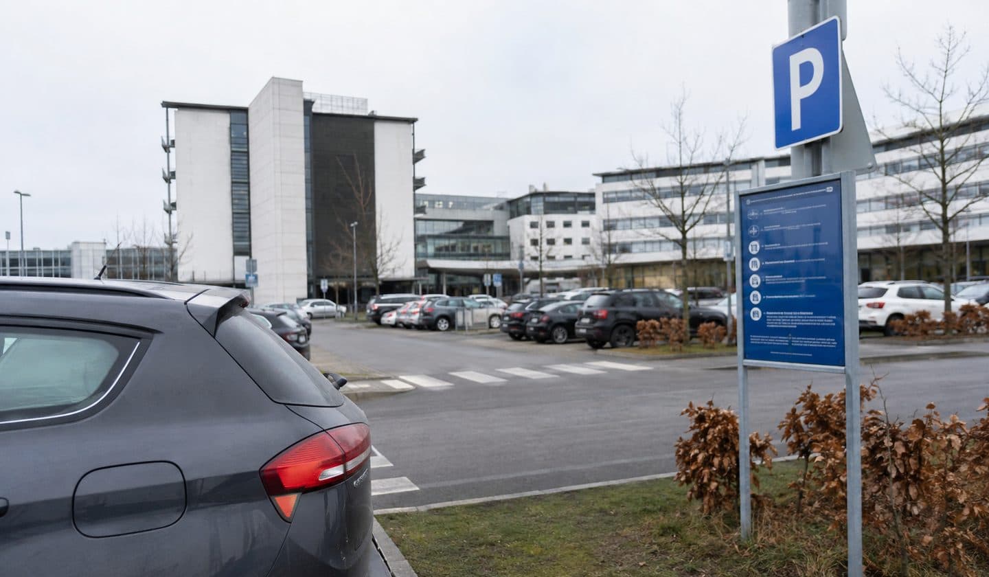 Parkeringsplads ved et dansk hospital med blåt p-skilt og infotavle ved indkørslen.